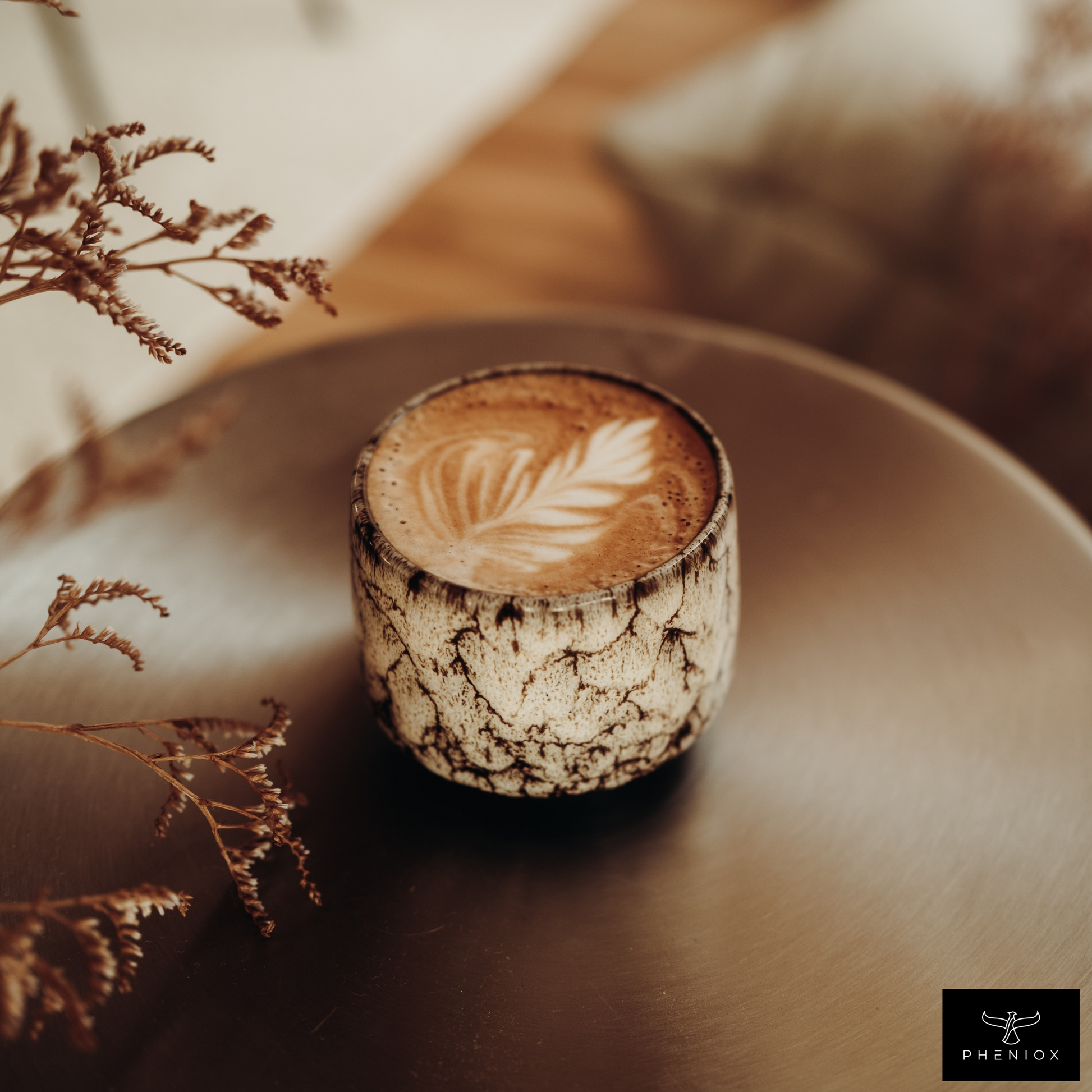 weiße Cappuccino Tasse mit Latte Art steht auf Metalltisch. Pflanze ragt im Vordergrund ins Bild