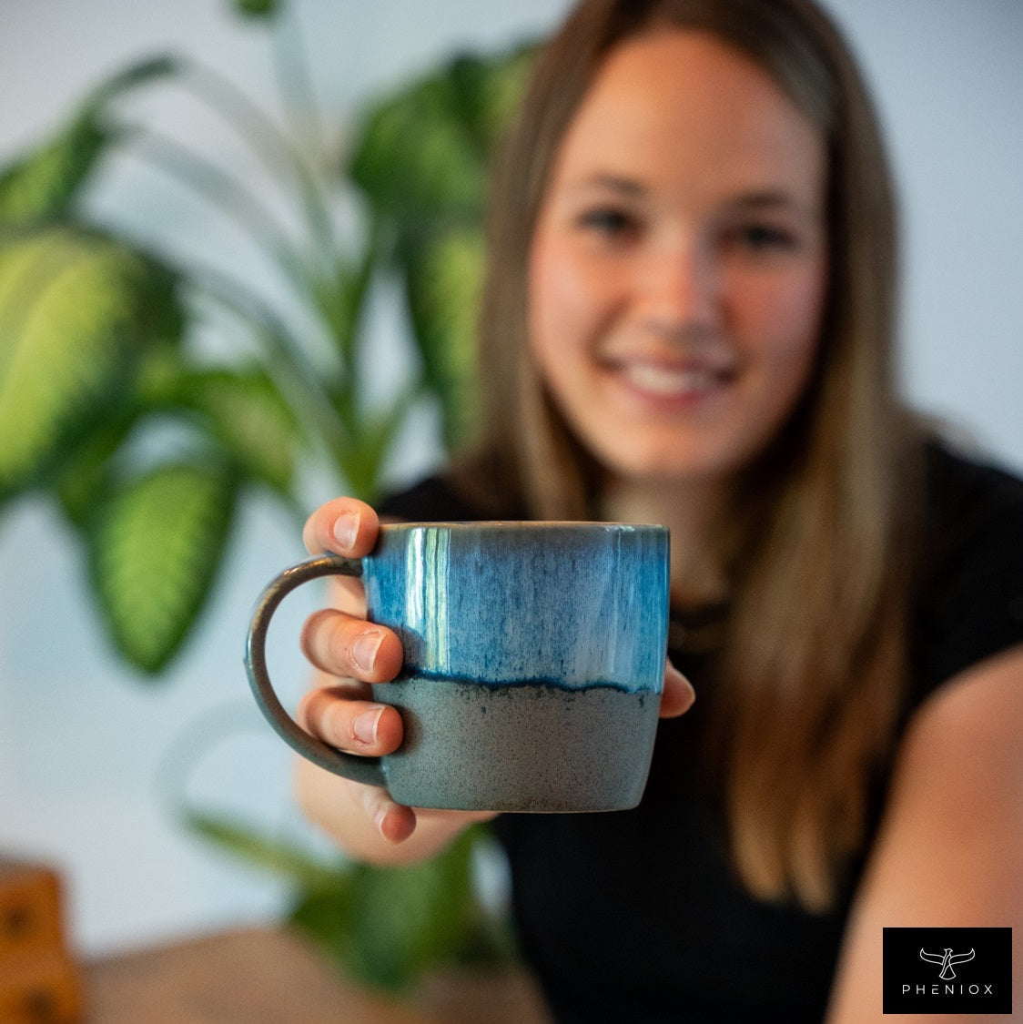 Der Fokus liegt auf einer PHENIOX Ocean Cappuccino Tasse mit blauem Farbverlauf. Eine Frau hält die Tasse ins Bild. Grüne Pflanzen im Hintergrund.