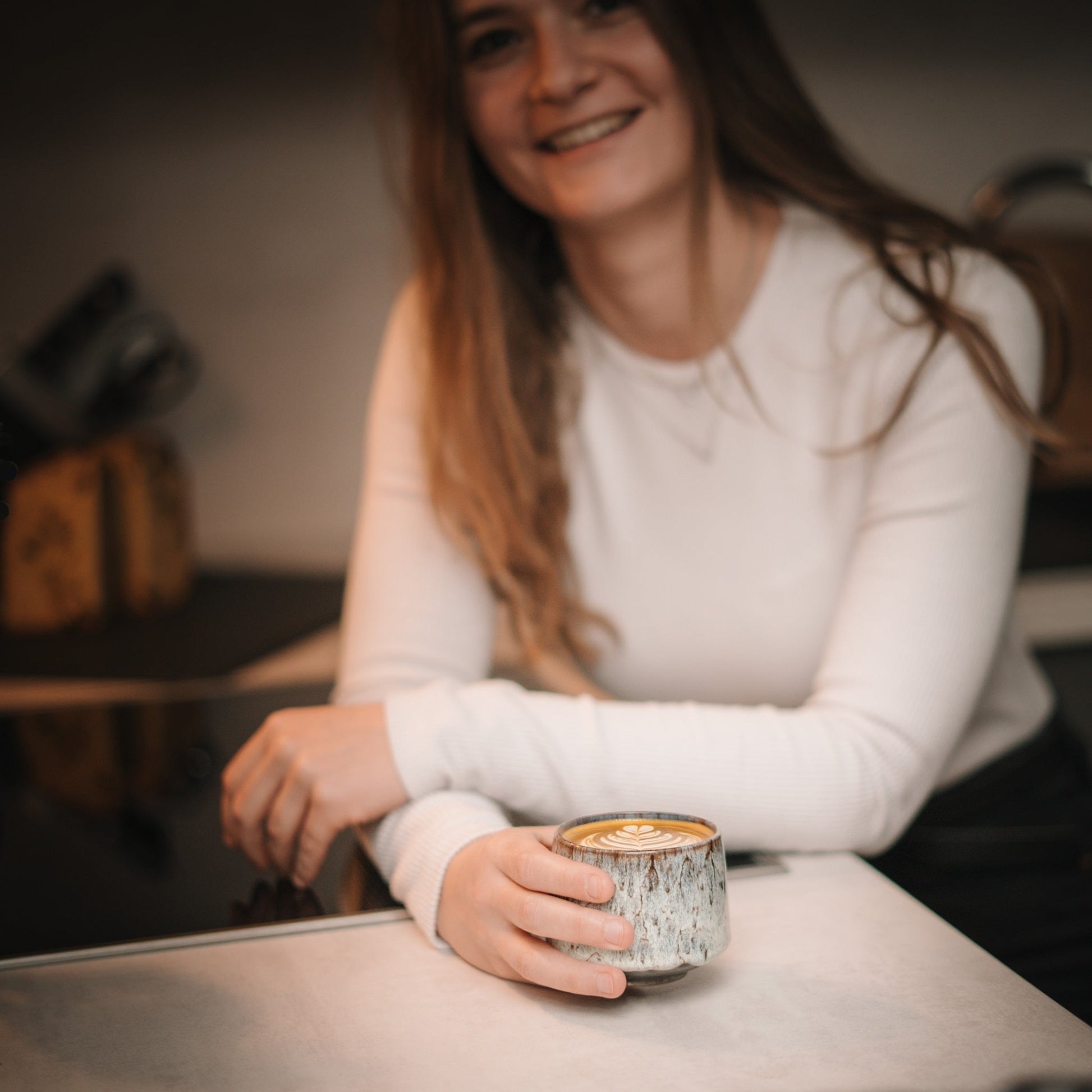 Frau lächelt in die Kamera und hält PHENIOX Cappuccino Tasse Tibet Misty mit Latte Art in der Hand.