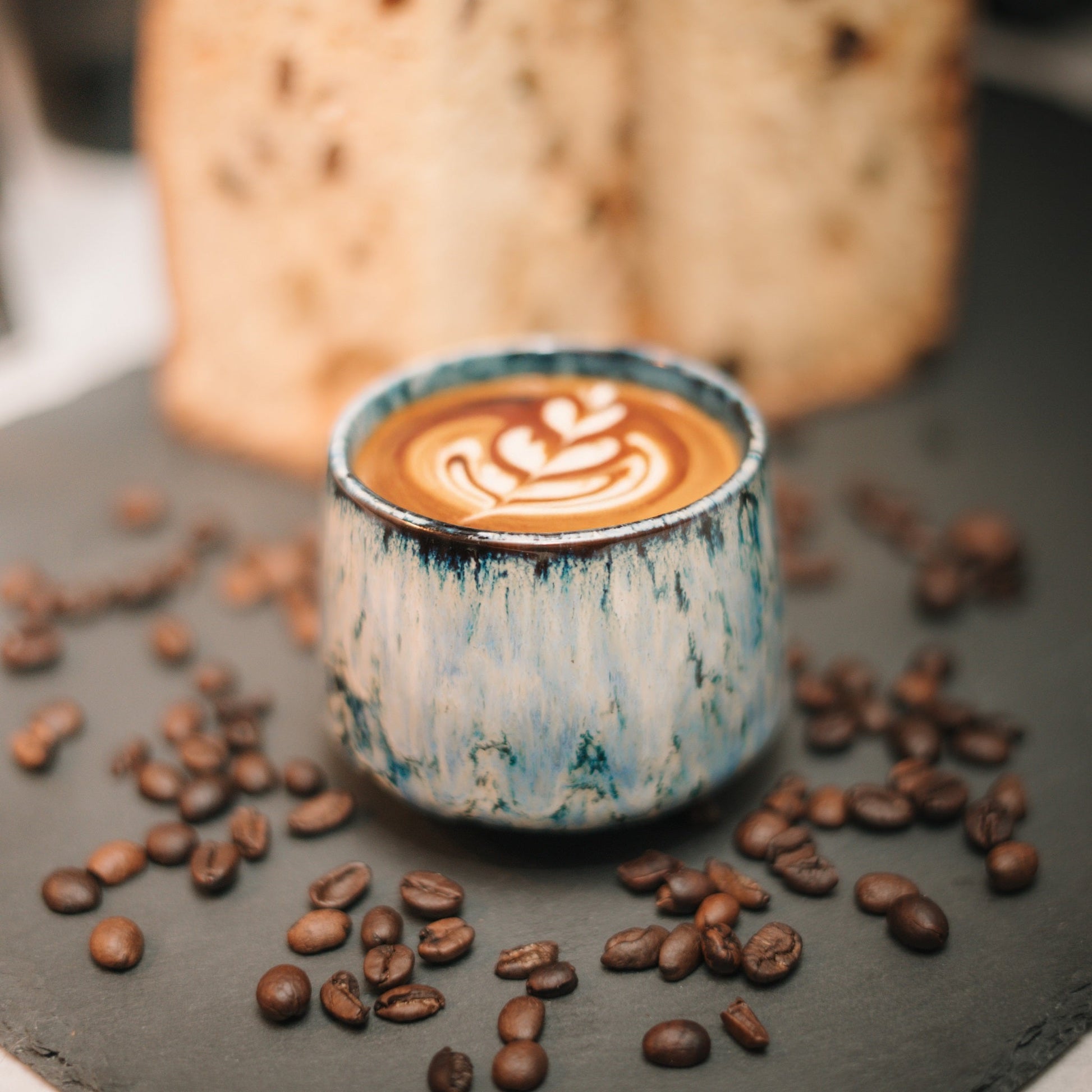 Blau-weiße Cappuccino Tasse mit Latte Art vor einem Kuchen mit Kaffeebohnen daneben.
