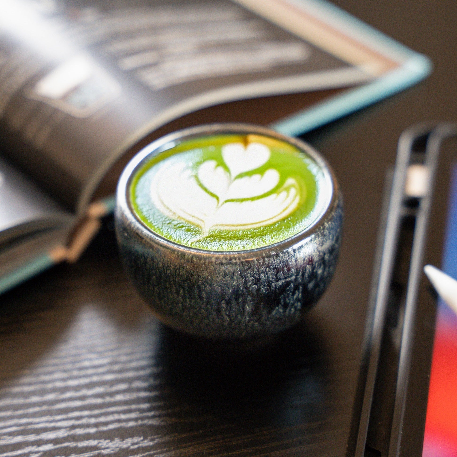 PHENIOX Espressotasse Galaxy steht auf einem schwarzen Tisch zwischen einem Tablet und einem Buch. In der Tasse ist Matche mit Latte Art.