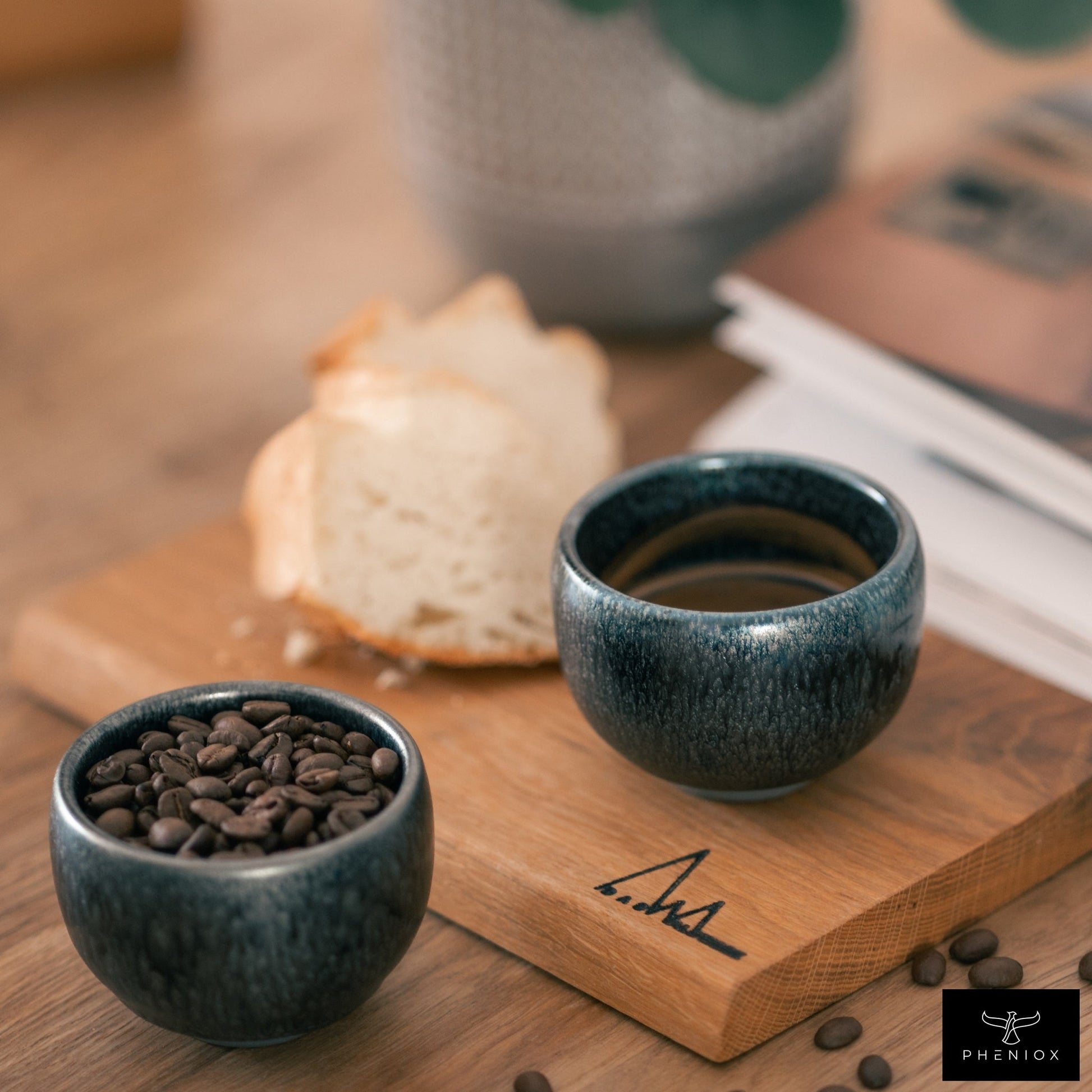 Zwei PHENIOX Espressotassen Galaxy aus Steingut stehen auf einem Tisch. Eine leicht erhöht auf einem Brettchen mit einem Stück Kuchen im Hintergrund. Eine Tasse ist mit einem Kaffee gefüllt, die andere mit Kaffeebohnen.