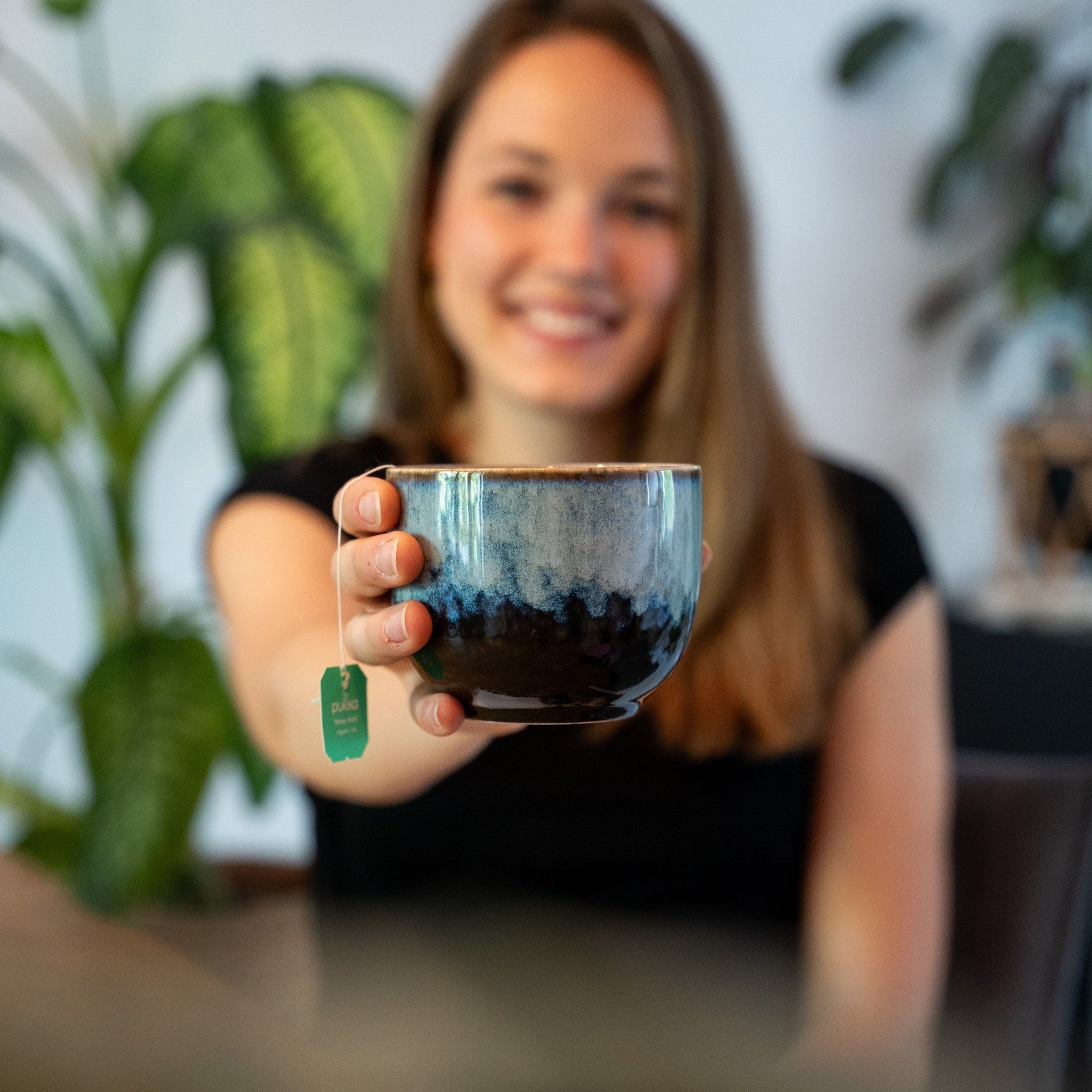 Junge Frau hält die PHENIOX Teetasse Infinity XL in einem schönen blau-schwarz in die Kamera. Im Hintergrund sind gründe Pflanzen zu sehen.