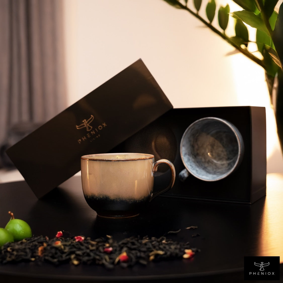 PHENIOX Teetassen Infinity XL stehen dekorativ auf einem schwarzen Holztisch zusammen mit der PHENIOX Geschenkbox. Im Vordergrund liegt etwas Tee auf dem Tisch.